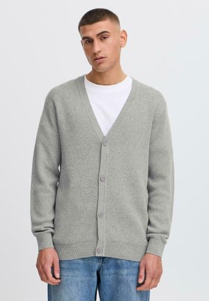 Šedý pletený cardigan s výstřihem do V, se pěti knoflíky, žebrovanými manžetami a spodním okrajem, nošený přes bílou košili, v kombinaci s modrými džíny.