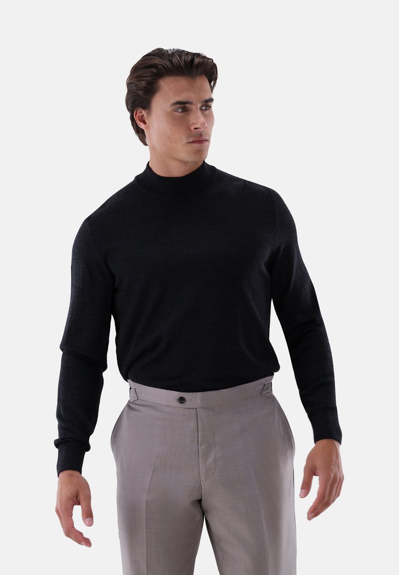 Mann steht da, trägt einen schwarzen Langarmrollkragenpullover und hellgraue maßgeschneiderte Hosen und schaut leicht zur Seite.