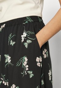 Jupe noire avec motif floral comprenant des fleurs roses et des papillons verts. Taille élastique et détail de poche latérale visibles. Texture de tissu douce.