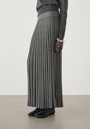 Osoba o ciemnej karnacji ubrana w grafitową plisowaną spódnicę, pasujący sweter, czarne buty oraz masywną srebrną bransoletkę, stojąca na gładkiej podłodze.