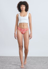 Bílý zkrácený top a červené spodní plavky s bílým ručně psaným vzorem. Model stojící na světlém šedém pozadí.