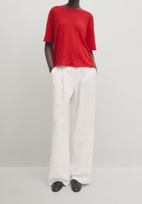 T-shirt rouge à manches courtes en tissu lisse, associé à un pantalon blanc taille haute et à jambes larges ; des chaussures noires élégantes complètent la tenue.