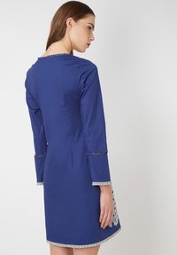 Vestido azul rey con mangas largas, dobladillo texturizado y delicados acentos de encaje en los puños. La vista posterior muestra una silueta ajustada y un detalle mínimo.