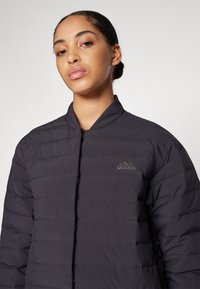 Juodas, siuvinėtas pūkinis kailis su stovinčiu apykakle ir priekiniais užtrauktukais. Kairėje krūtinės srityje yra subtilus, toninis adidas logotipas.
