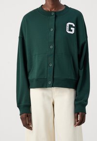 Grüner cropped Sweatshirt mit Knopfverschluss, zwei Fronttaschen und einem weißen "G"-Emblem, das auf der Brust gestickt ist. Weiches Material mit langen Ärmeln.