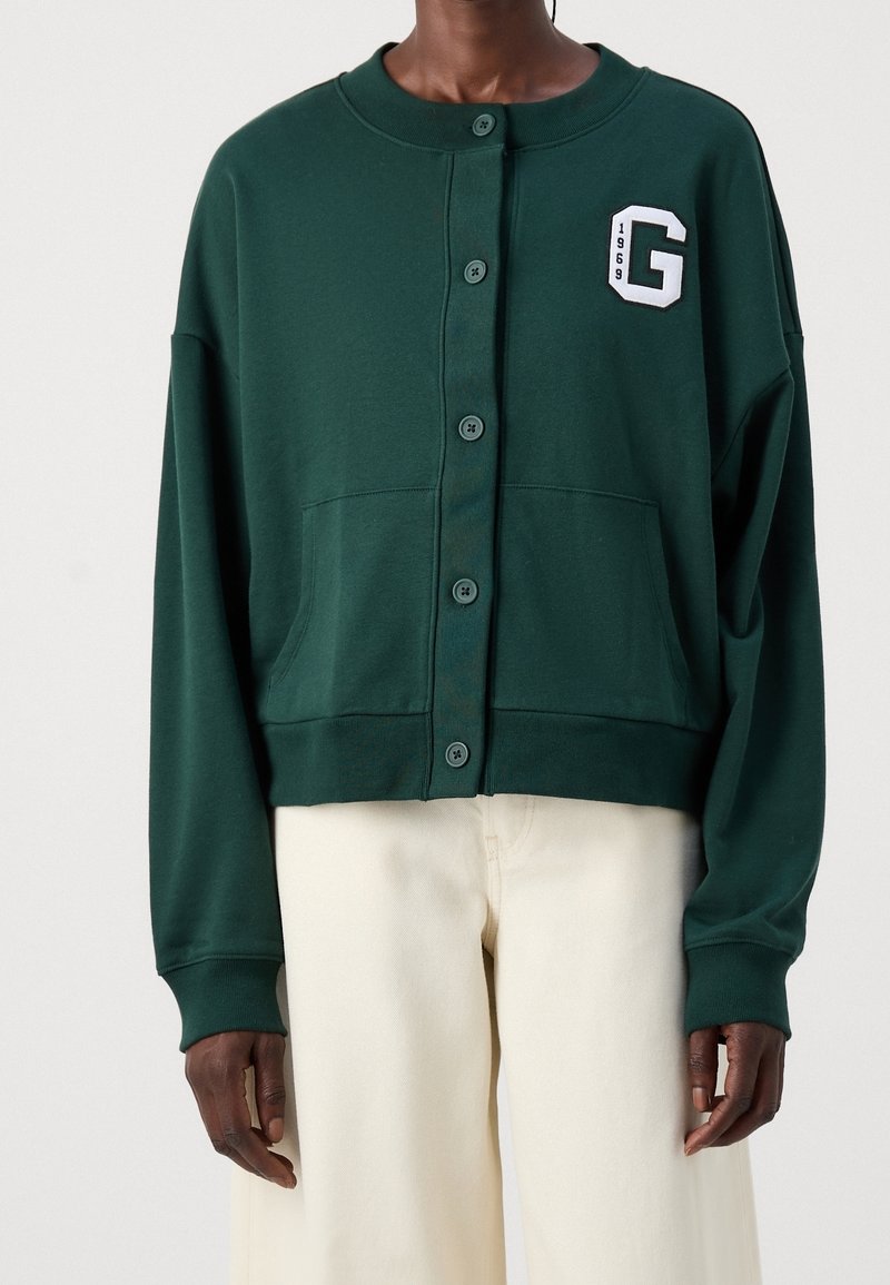 Grüner cropped Sweatshirt mit Knopfverschluss, zwei Fronttaschen und einem weißen "G"-Emblem, das auf der Brust gestickt ist. Weiches Material mit langen Ärmeln.