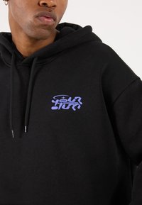 Black hoodie made from soft fabric, featuring a purple graphic logo on the chest, and finished with a kangaroo pocket and drawstring hood.