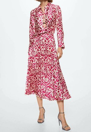 Femme portant une robe midi à manches longues rose à motifs avec un col, et des sandales à talons hauts noires, debout devant un fond blanc.