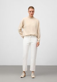 Beżowy sweter z dzianiny z wysokim kołnierzem, zestawiony z białymi, prostymi dżinsami i beżowymi trampkami. Prosty fason, miękka tekstura.