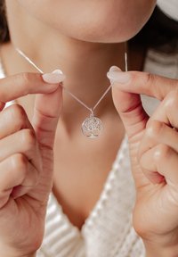 Ciondolo a forma di albero della vita in argento su una catena sottile, con intricate fessure che rappresentano rami e foglie, tenuto tra le dita.