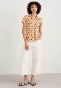 Krátky rukáv oranžovej košele s bielym a tmavomodrým vzorom slnečnica, v kombinácii s krémovými širokými nohavicami a šedými sandálmi, stojacou na neutrálnom pozadí.