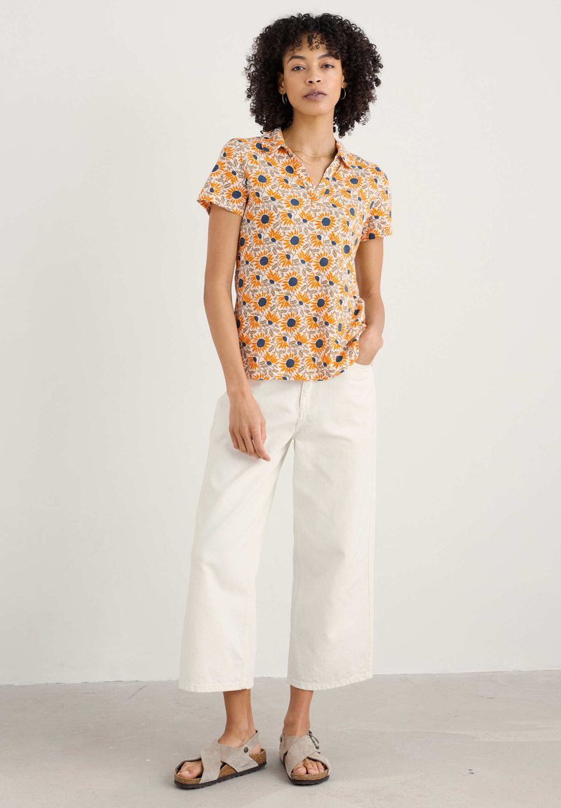 Krátky rukáv oranžovej košele s bielym a tmavomodrým vzorom slnečnica, v kombinácii s krémovými širokými nohavicami a šedými sandálmi, stojacou na neutrálnom pozadí.
