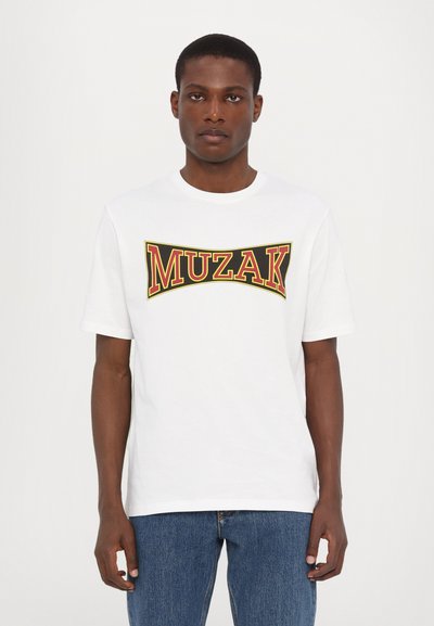 T-shirt en coton blanc avec un graphisme "MUZAK" audacieux en rouge et jaune. Manches courtes, col rond, coupe standard. Design décontracté avec une texture lisse.