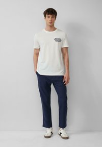 Witte katoenen t-shirt met marineblauw grafisch print, gecombineerd met marineblauwe broek. Model draagt witte sneakers met zwarte en bruine accenten.