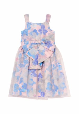 Junges Mädchen trägt ein pastellfarbenes Blumenkleid mit einer großen Schleife, hält den Stoff und steht vor einem neutralen Hintergrund.