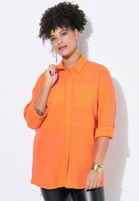Camicia button-up di un vivace arancione con tessuto testurizzato, maniche arrotolabili e due tasche sul petto. Accompagnata da dettagli in oro.