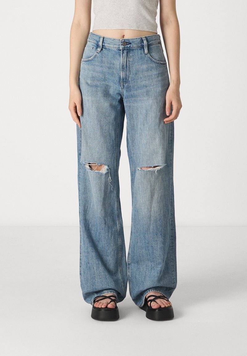 Personne portant un jean large bleu clair avec des déchirures aux genoux et des sandales compensées noires, debout devant un fond blanc uni.