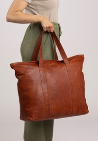 Grote bruin leren tote bag met twee stevige handvatten, een gladde textuur en subtiele naaddetails. Geplaatst op taillehoogte tegen een grijze achtergrond.