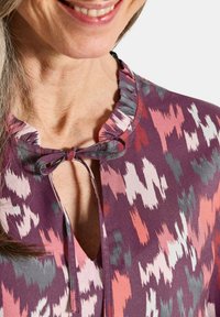 Lila Bluse mit einem VOLANT-Ausschnitt und einem Bindedetail. Mit pinken, grauen und weißen abstrakten Formen auf einem strukturierten Stoff bedruckt.