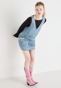 Denim overall dress with a deep V-neck, two zippered pockets, and a faded blue hue, paired with knee-high pink cowboy boots.