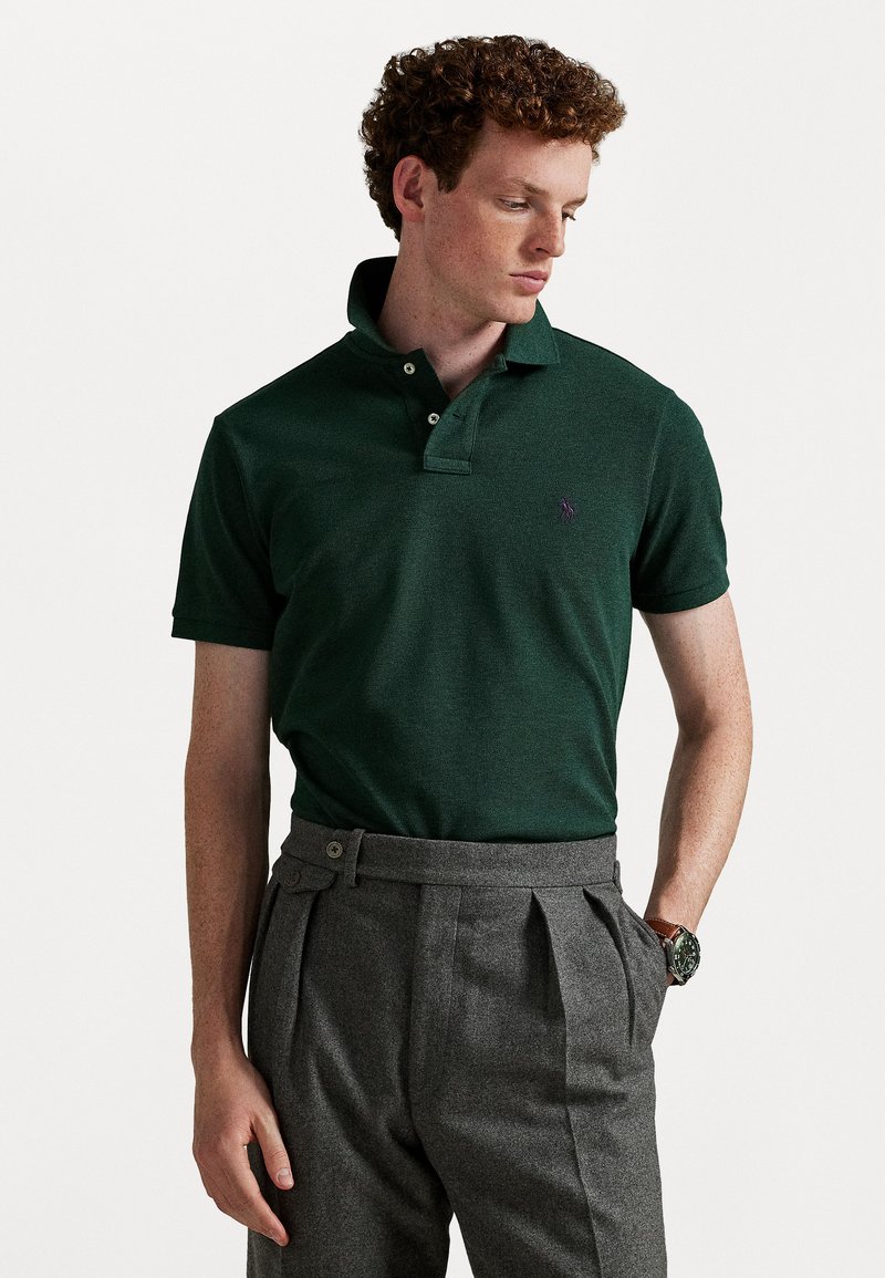 Tricou polo din bumbac verde închis, cu un gât cu trei butoane și un mic logo mov. Asortat cu pantaloni gri plisati și un ceas.