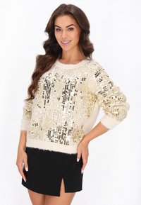 Beige pulóver, arany és ezüst flitterekkel díszítve, kerek nyakkivágással és laza fazonnal, fekete szoknyával párosítva, amely oldalhasítással rendelkezik.