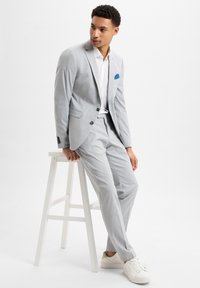 Lichtgrijs pak met slim fit ontwerp, met zwarte knopen en een blauwe pochette. Getextureerde stof, gecombineerd met een wit overhemd en sneakers.