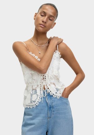 Witte kanten top met bloemenpatronen, verstelbare strikken in de taille en golvende randen, gecombineerd met lichtblauwe high-waisted denim broeken.
