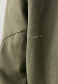 Oliven grøn sweatshirt med en blød tekstur. Har en ribbet halsudskæring og teksten "Scispiritual" trykt i lys grøn på ærmet.