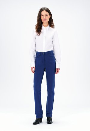 Witte knoopsluiting overhemd en blauwe slim-fit broek met voorkanten. De broek heeft riemlussen. Model draagt zwarte hakken.