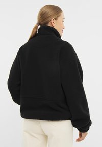 Pullover in pile nero con colletto alto, spalle scese e maniche lunghe. Tessuto morbido con una vestibilità rilassata, accentuato da una linea di cucitura bassa.