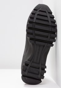 Černá podrážka s hlubokým vzorem a vystouplým nápisem "Calvin Klein Jeans" ve středu, nakloněná na pozadí s bílými rohy.