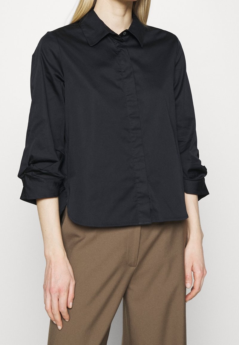 Femme portant une chemise noire à manches longues boutonnée avec les manches retroussées et un pantalon marron, debout devant un fond uni.