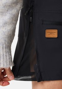 Svart utomhusjacka med dragkedjeficka, nätfoder och läderetikett. Hand mot tyget visar materialets textur.