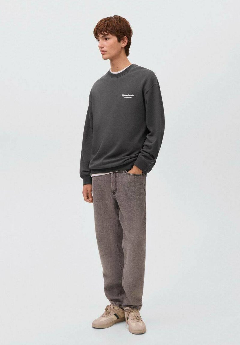 Felpa grigia scura con scollo a girocollo e dettagli del logo bianco, abbinata a jeans grigi larghi e sneaker di colore chiaro.