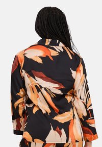 Veste noire avec un imprimé floral multicolore en orange et crème, dotée de manches longues et d'un col classique, en tissu lisse.