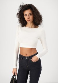 Ženska s kodrastimi lasmi, oblečena v beli dolg rokav in kratko majico ter temne kavbojke, drži črno torbico v eni roki.