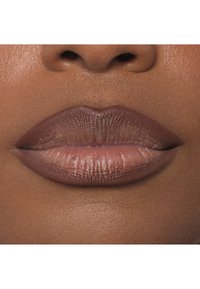 Zbliżenie pełnych ust pokrytych matową, cielistą szminką, o gładkiej fakturze i lekki połysku na dolnej wardze, na tle ciemnej skóry.