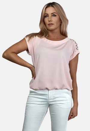 Jasnoróżowa bluzka z krótkim rękawem, okrągłym dekoltem i koronkowymi detalami na ramionach, zestawiona z białymi spodniami o slim-fit. Gładka tekstura materiału.