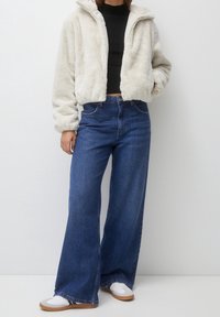 Veste courte en fausse fourrure crème avec capuche, associée à un jean bleu taille haute à jambes larges. Baskets blanches avec semelles marron.