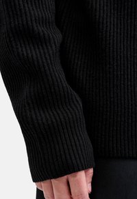 Schwarzer gerippter Pullover mit strukturiertem Strick, in lockerer Passform und mit breiten Bündchen, vor einem neutralen Hintergrund präsentiert.