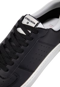 Schwarze Ledersneaker mit weißer Gummisohle, runder Zehenform, perforierten Details, schwarzen Schnürsenkeln und einem Markenlabel auf der Zunge.