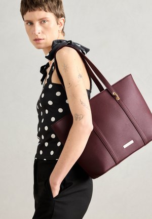 Burgunder farget lær tote bag med strukturert overflate, detaljer i gull, kombinert med en svart og hvit prikkete topp, modell som holder vesken i armen.