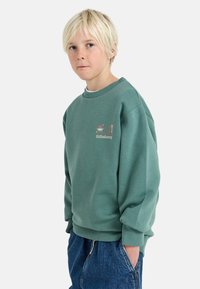 Sudadera de forro polar verde con cuello redondo y puños acanalados, que presenta un pequeño logotipo en el pecho y un ajuste relajado. Combinada con pantalones cortos de mezclilla azules.