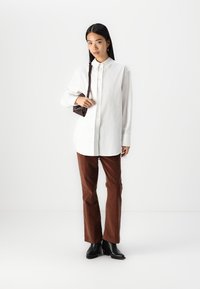 White button-up shirt, brown corduroy pants, black heeled boots. Model holding a small dark brown bag, standing against a plain background.