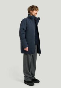 Mörkblå vadderad jacka med hög krage, försedd med full dragkedja och en något löst passform. Matcher med grå skräddarsydda byxor och svarta skor.