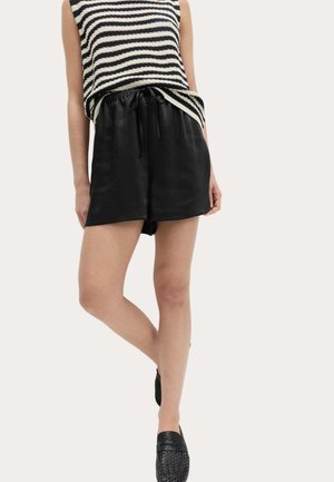 Kvinde iført sorte shorts med snor, sorte vævede slip-on sko og en ærmeløs sort-hvidstripet top, stående med benene krydsede.
