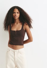 Žena s dlhými kučeravými vlasmi má na sebe bezramienkový hnedý crop top a svetlohnedé nohavice s vysokým pásom, stojí pred jednoduchým bielym pozadím.