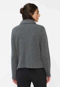 Donna con capelli scuri raccolti in un chignon, che indossa un blazer grigio a zigzag e pantaloni neri, rivolta leggermente di lato su uno sfondo bianco.