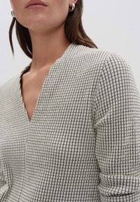 Donna che indossa un top cremoso a trama con un motivo a quadri neri, scollo a V e maniche arrotolate, mostrata dalla mascella fino a metà torso.